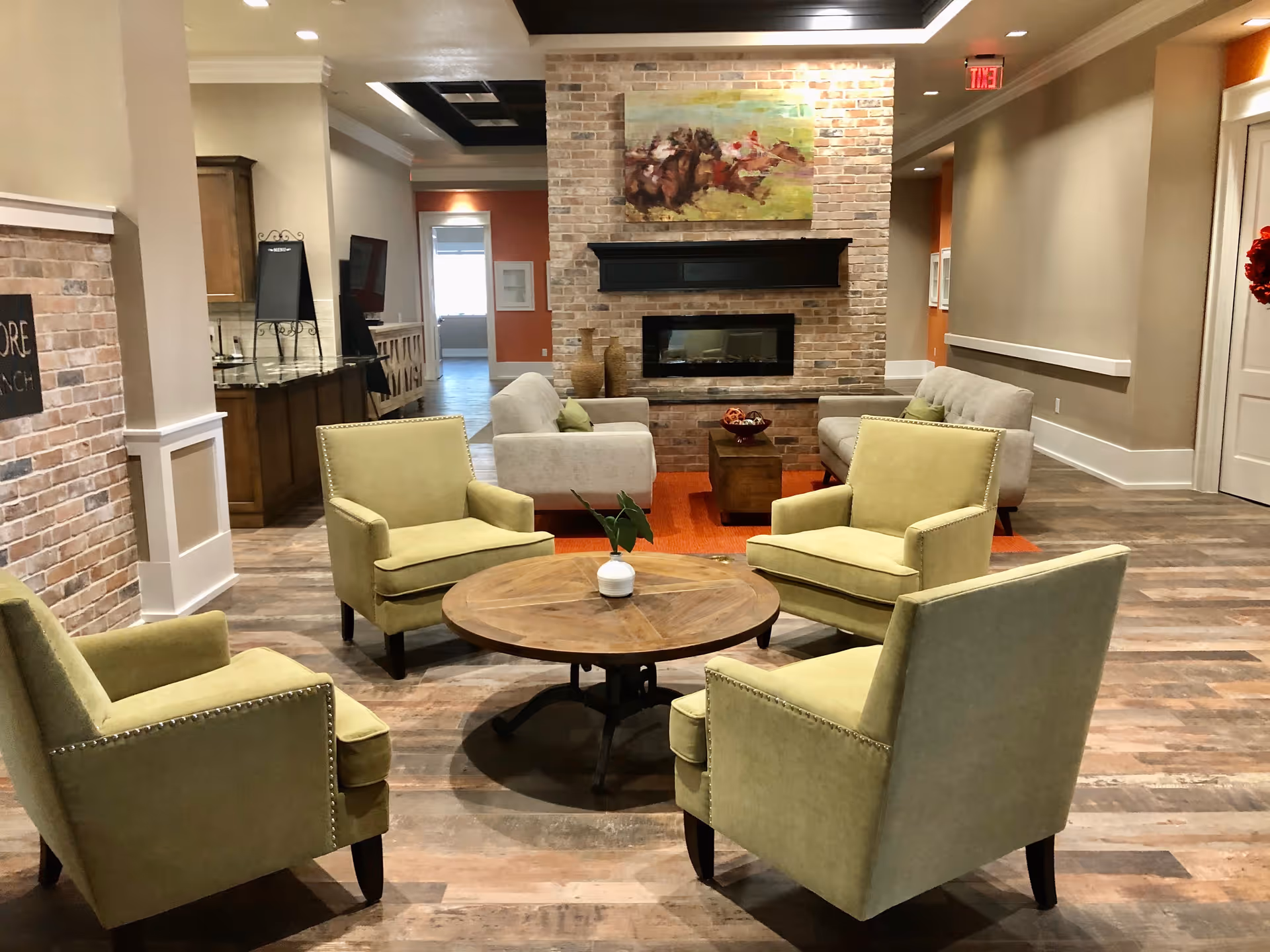 Cozy communal lounge with four green armchairs around a round wooden table facing a brick fireplace and sofas.
