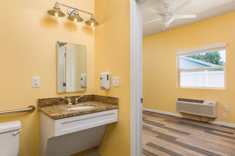Yellow-walled interior showing a bathroom sink with granite countertop, mirror and grab bar, opening into a room with wood-look floors, window, ceiling fan and wall air unit.