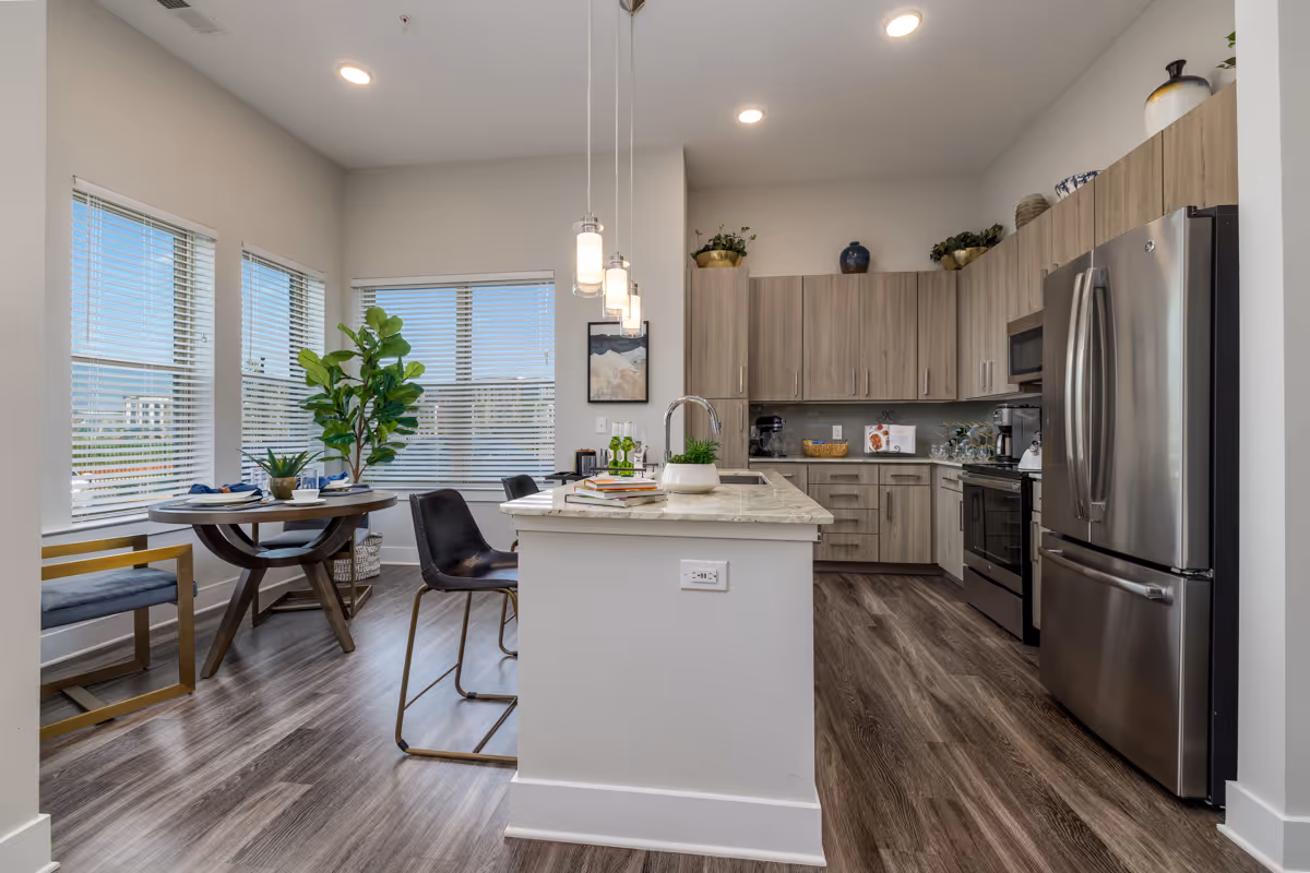 Modern open kitchen with an island, stainless steel refrigerator, wood cabinets, and a small dining table by bright windows.