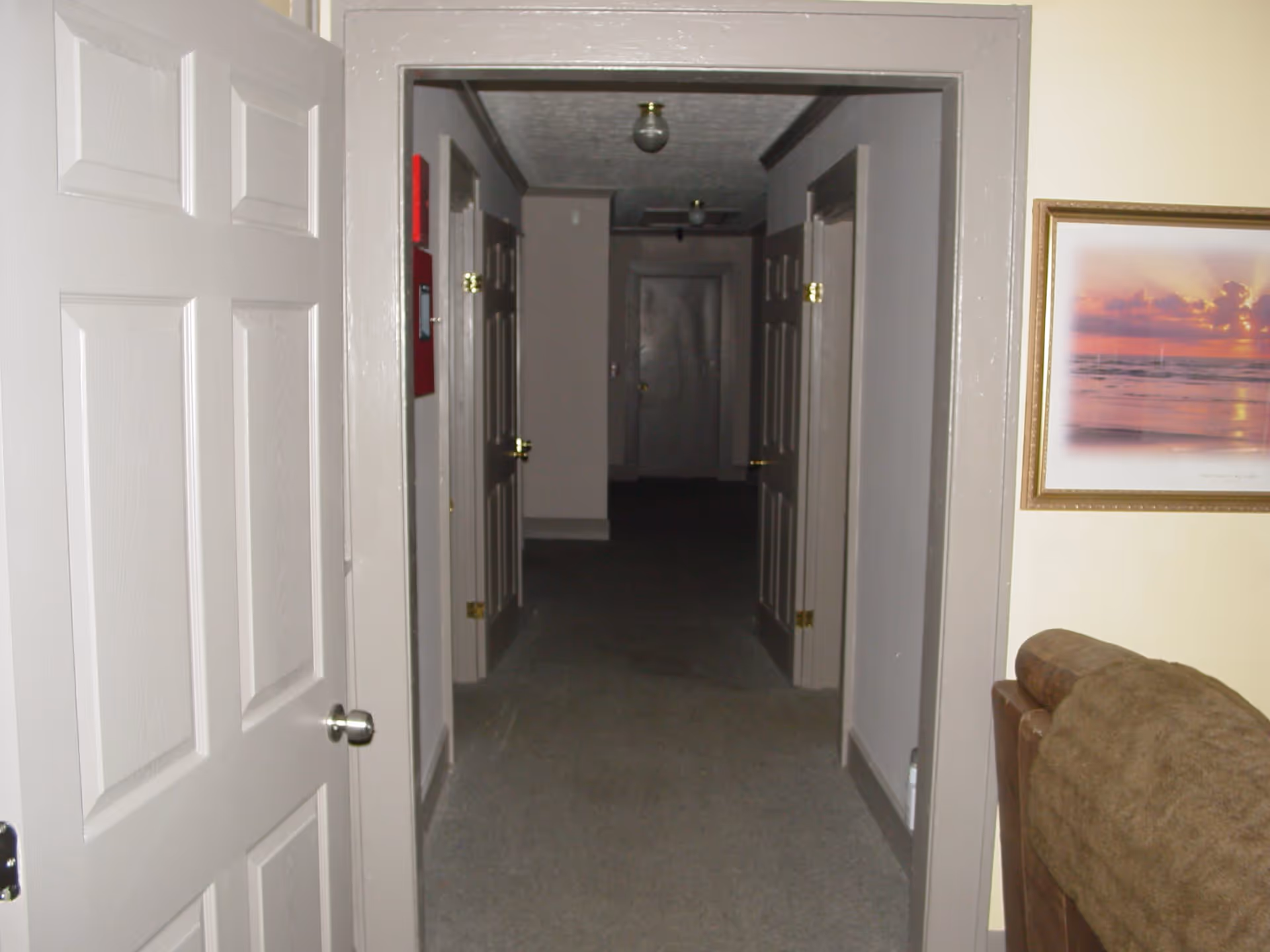View of a hallway in a facility with several closed doors on both sides, beige walls, and carpeted floor. A partially visible brown couch and a framed picture of a sunset over the ocean are on the right side near the hallway entrance.
