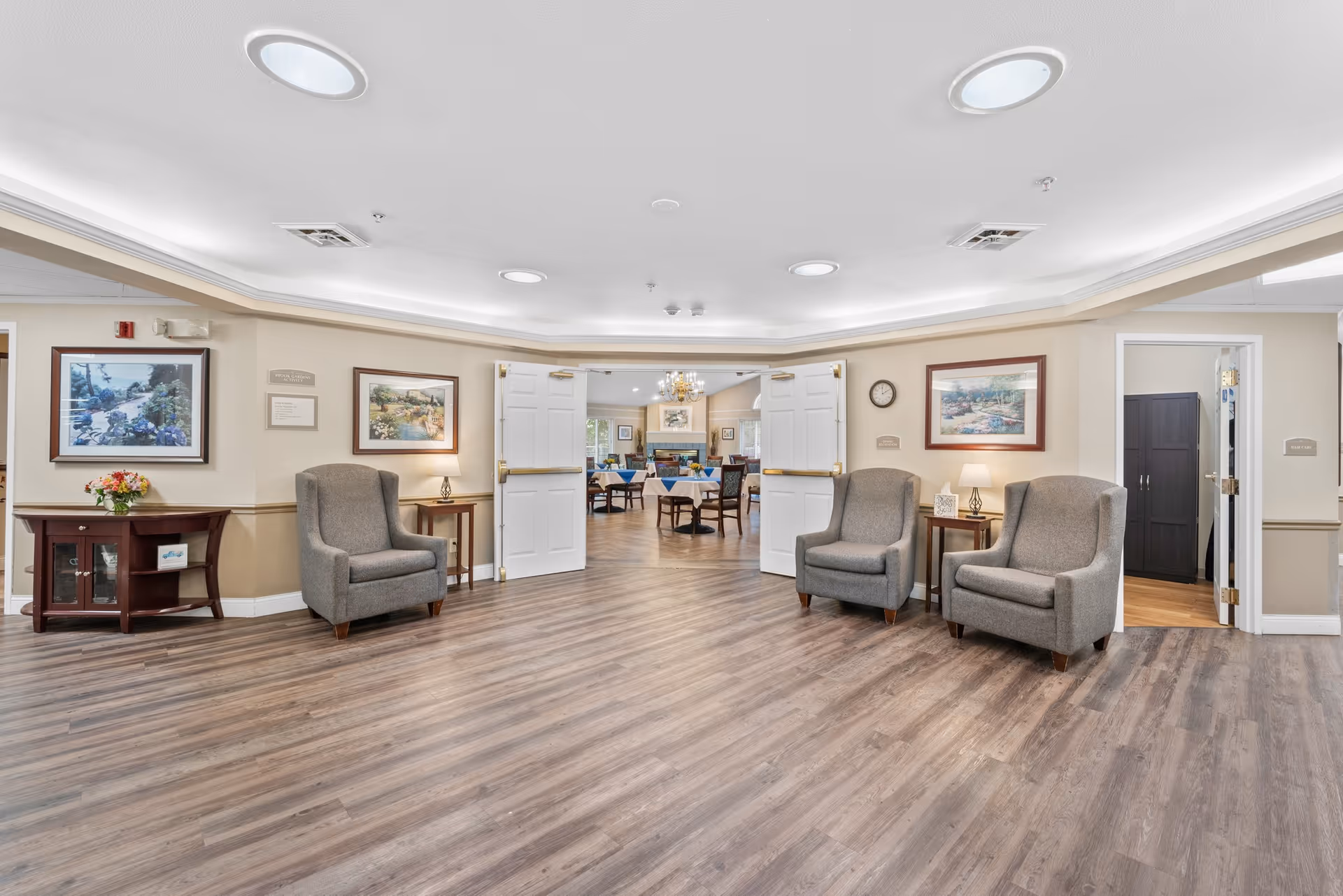 A spacious, well-lit common area with four gray armchairs arranged along the walls, two small side tables with lamps, framed landscape paintings, and double white doors leading to a dining room with tables and chairs. The floor is wood laminate, and the ceiling has recessed lighting.