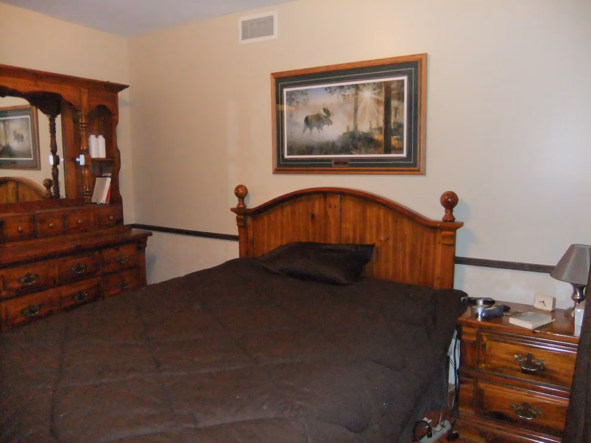 A bedroom with a wooden bed frame and matching wooden dresser and nightstand. The bed is covered with a dark brown comforter and has a single pillow. Above the bed hangs a framed picture of a moose in a forest with sunlight streaming through the trees. The nightstand has a lamp, a book, and some electronic devices on it.