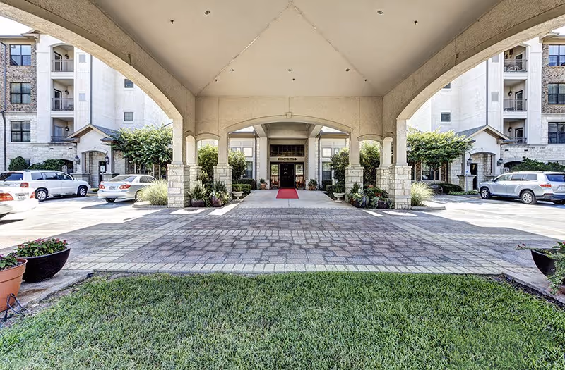 Covered entrance area of Conservatory At Keller Town Center with stone pillars, potted plants, a red carpet leading to the main door, and parked cars on either side.