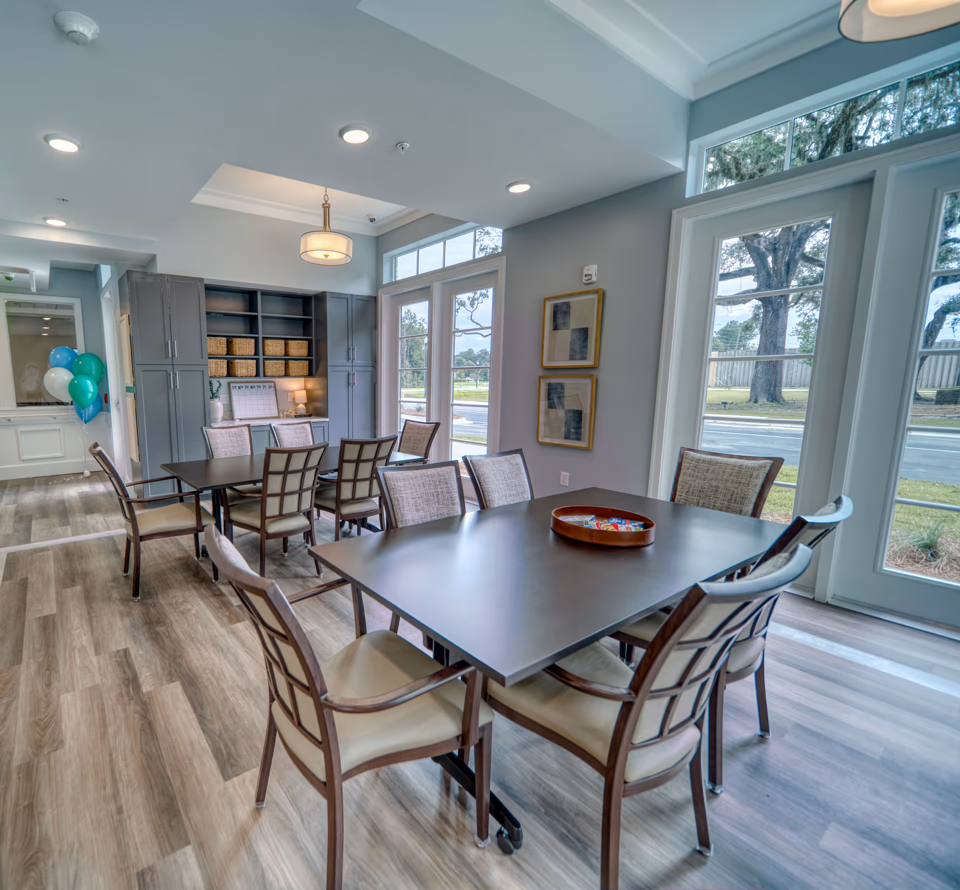 A bright and modern dining area in an assisted living facility with two dark wood tables surrounded by cushioned chairs. The room features large windows and glass doors letting in natural light, light wood flooring, gray walls, built-in cabinetry with baskets, and a ceiling light fixture. Balloons are visible in the background near a hallway.