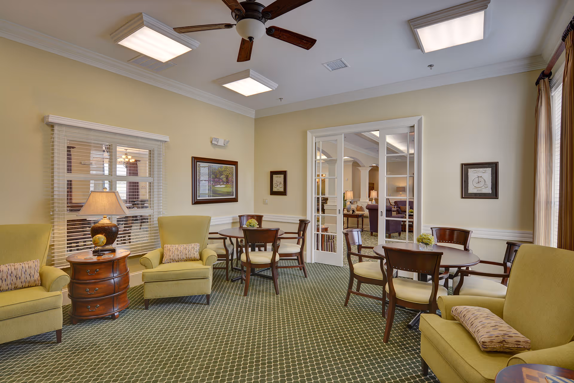 Bright communal sitting area with upholstered armchairs, round tables and chairs, side tables and a ceiling fan in a senior living facility.
