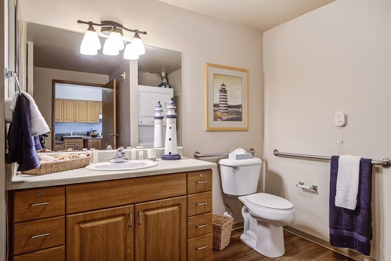 A bathroom with a wooden vanity featuring a sink and a large mirror above it. On the countertop are decorative lighthouse figurines, a basket with towels, and toiletries. The toilet is next to the vanity with grab bars on the walls for accessibility. A framed picture of a lighthouse hangs above the toilet. The floor is wood, and a towel rack with a white and purple towel is mounted on the wall.