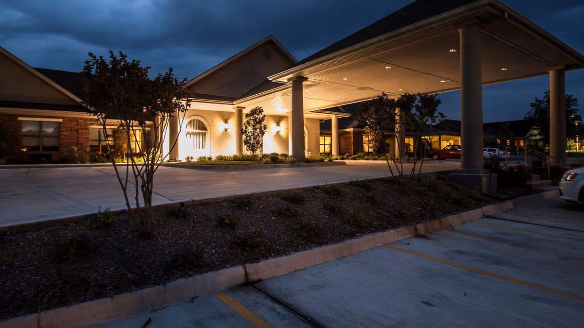 Night view of the front entrance of The Plaza at Twin Rivers facility, showing a covered drop-off area with columns, illuminated exterior walls, landscaped plants, and a parking area with cars.