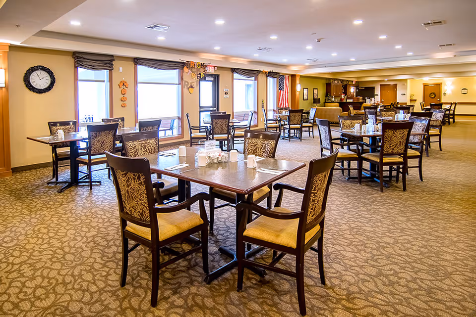 A spacious dining room in a senior living facility with multiple square tables and cushioned chairs arranged neatly. The room has large windows with brown valances, a wall clock, and an American flag near the back. The carpet has a patterned design, and the walls are painted in warm tones. The tables are set with white mugs, salt and pepper shakers, and napkin holders.
