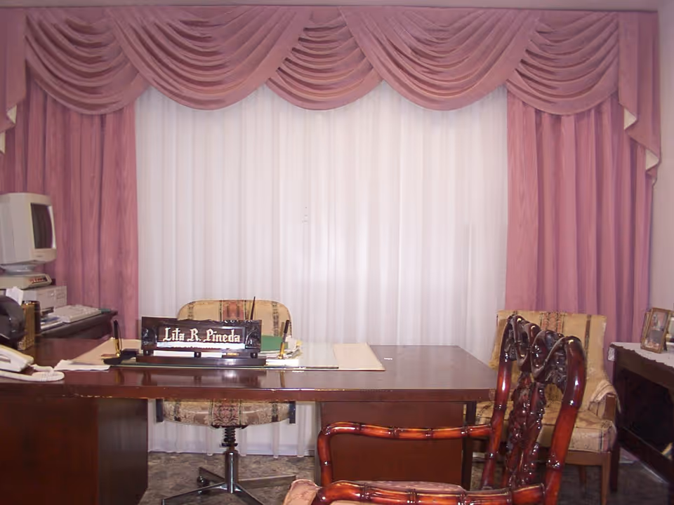 An office room with a large wooden desk in the center. Behind the desk is a chair with a patterned fabric. On the desk is a nameplate that reads 'Lita R. Pineda', along with some papers and office supplies. To the left, there is an old computer monitor and keyboard. The background features large windows covered with white sheer curtains and pink draped valances. There are two wooden chairs with carved backs and cushioned seats in front of the desk, and another cushioned chair to the right side of the room.