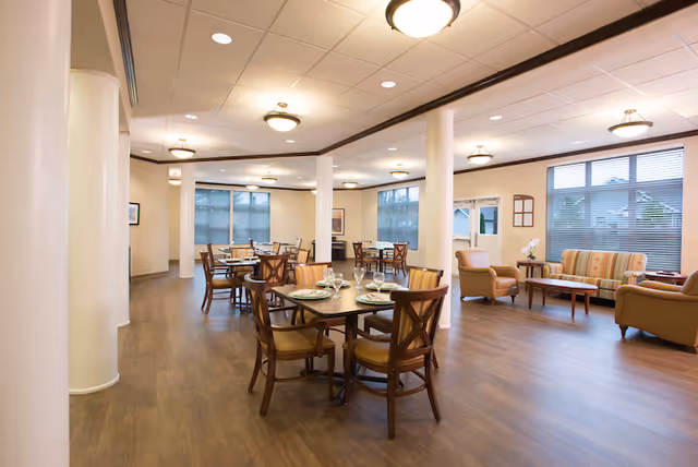 A spacious, well-lit dining area in a senior living facility with several wooden tables and chairs arranged neatly. The tables are set with plates, glasses, and silverware. There are large windows with blinds allowing natural light to enter. In the background, there is a seating area with a striped sofa, armchairs, and a coffee table. The room has wooden flooring, white walls, and ceiling lights.