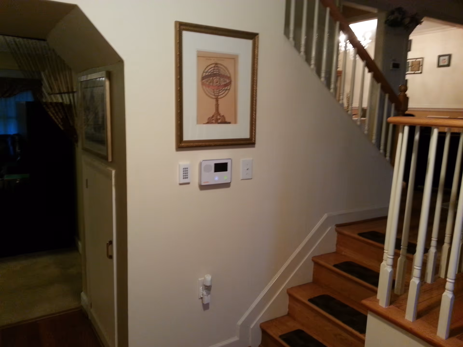 Interior view of a home showing a staircase with wooden steps and white railings. On the wall next to the stairs, there is a framed picture above a thermostat and light switches. To the left, there is a small door under an archway leading to a darker room with curtains.
