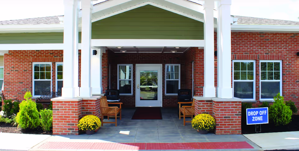 Entrance of a brick building with a covered porch supported by white columns. There are two wooden benches on either side of the entrance door, yellow potted flowers near the columns, and a blue sign on the right side that reads 'DROP OFF ZONE'.
