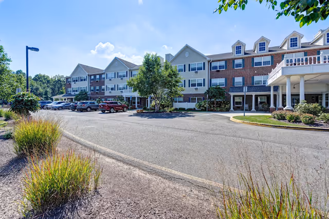Front exterior of a three-story brick-and-siding senior living building with a circular driveway, parked cars, and landscaping under a blue sky.