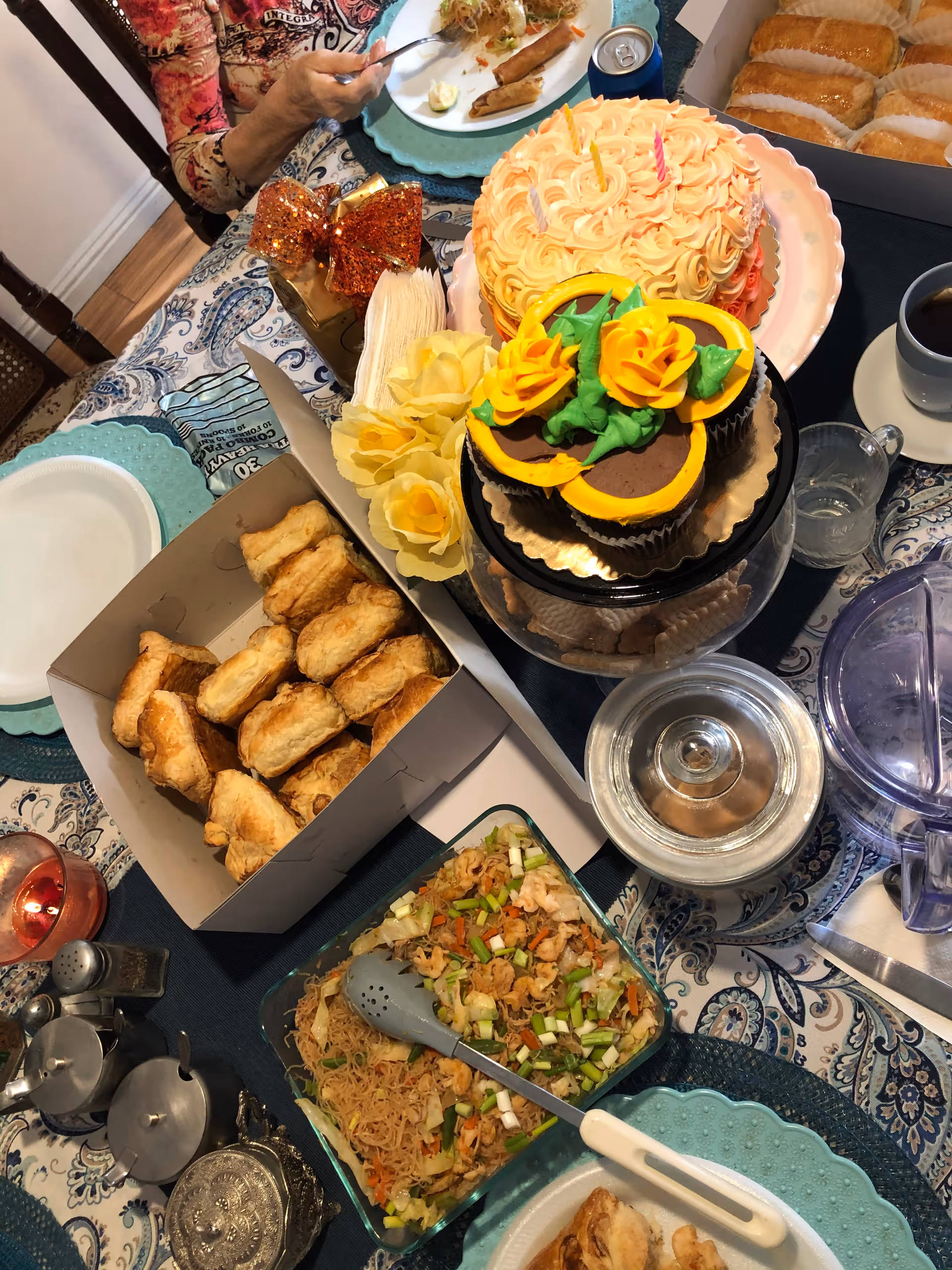 A dining table set with various foods including a box of pastries, a dish of stir-fried noodles with vegetables and shrimp, a decorated cake with yellow and orange frosting flowers, a pink frosted cake with a candle, a cup of coffee, a glass of water, and a water pitcher. A person is partially visible eating from a plate at the top left corner of the image.
