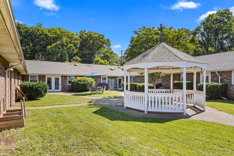 Outdoor courtyard area of Woods Cove Assisted Living featuring a white gazebo with seating inside, surrounded by green grass and bushes. The courtyard is enclosed by single-story brick buildings with windows and doors. There is a table with chairs and an umbrella in the background, and trees with green foliage under a blue sky with some clouds.