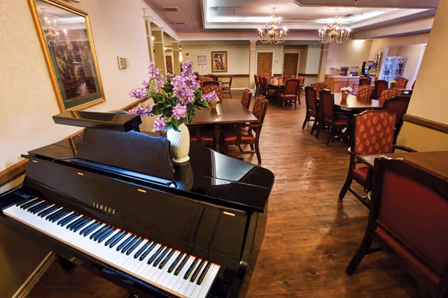 Interior view of a senior living facility dining room with multiple wooden tables and red cushioned chairs. A black Yamaha piano with a vase of purple flowers on top is in the foreground. The room has wooden flooring, chandeliers, framed artwork on the walls, and a warm, inviting atmosphere.