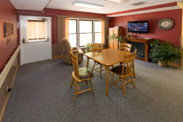 A cozy common area with a wooden table and four chairs in the center, a recliner chair near large windows with vertical blinds, a wall-mounted TV above a fireplace, a clock on the wall, and several potted plants. The walls are painted red and the floor is carpeted.