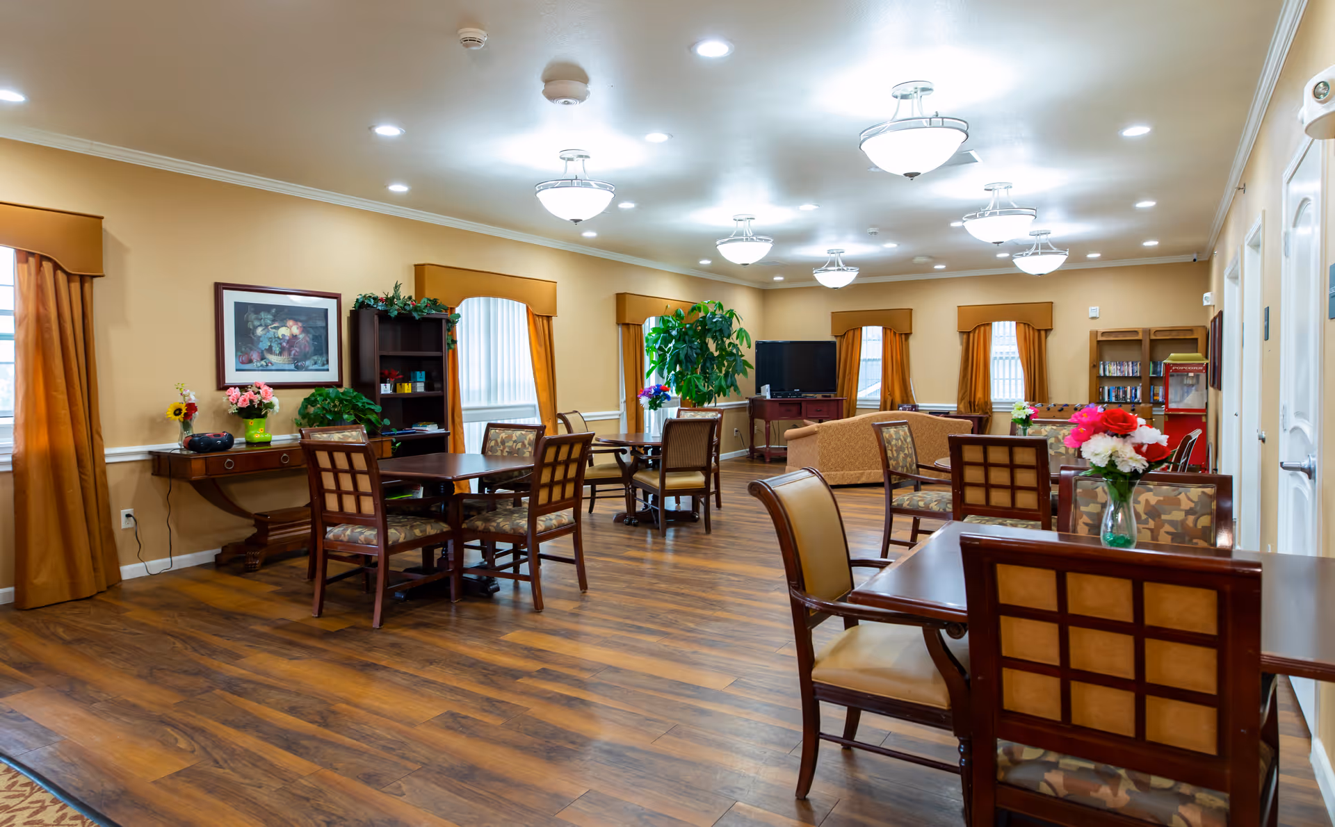 A spacious, well-lit common area in an assisted living facility featuring several wooden tables and chairs with patterned cushions. The room has warm beige walls, wood flooring, and multiple windows with gold curtains. There are decorative plants, flowers in vases on the tables, a bookshelf, a television, and a popcorn machine in the background.