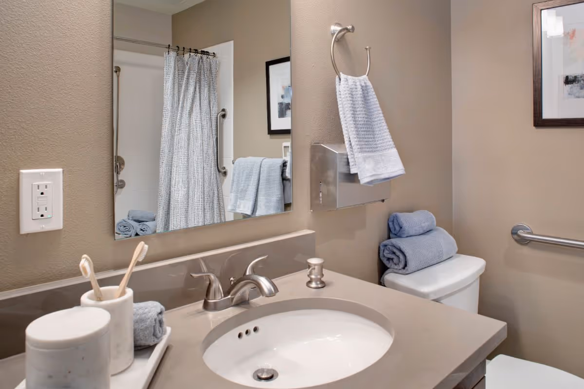 A clean and modern bathroom with a beige countertop and an oval sink. There are two toothbrushes in a holder, a small rolled towel, and a soap dispenser on the counter. A large mirror is mounted above the sink, reflecting a shower with a white curtain and grab bars. On the right side, there is a toilet with two folded blue towels on top, a towel ring with a white hand towel, and a framed picture on the wall.