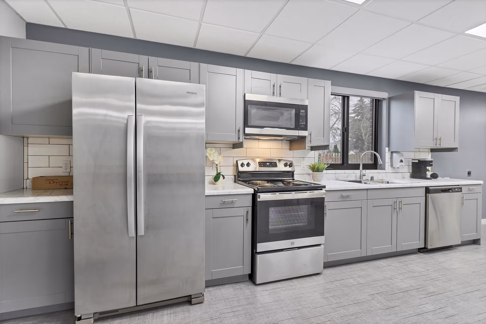 Modern kitchen with gray cabinets, stainless steel refrigerator, stove, microwave, dishwasher, and a sink under a window. The kitchen has a tiled backsplash and light-colored countertops.