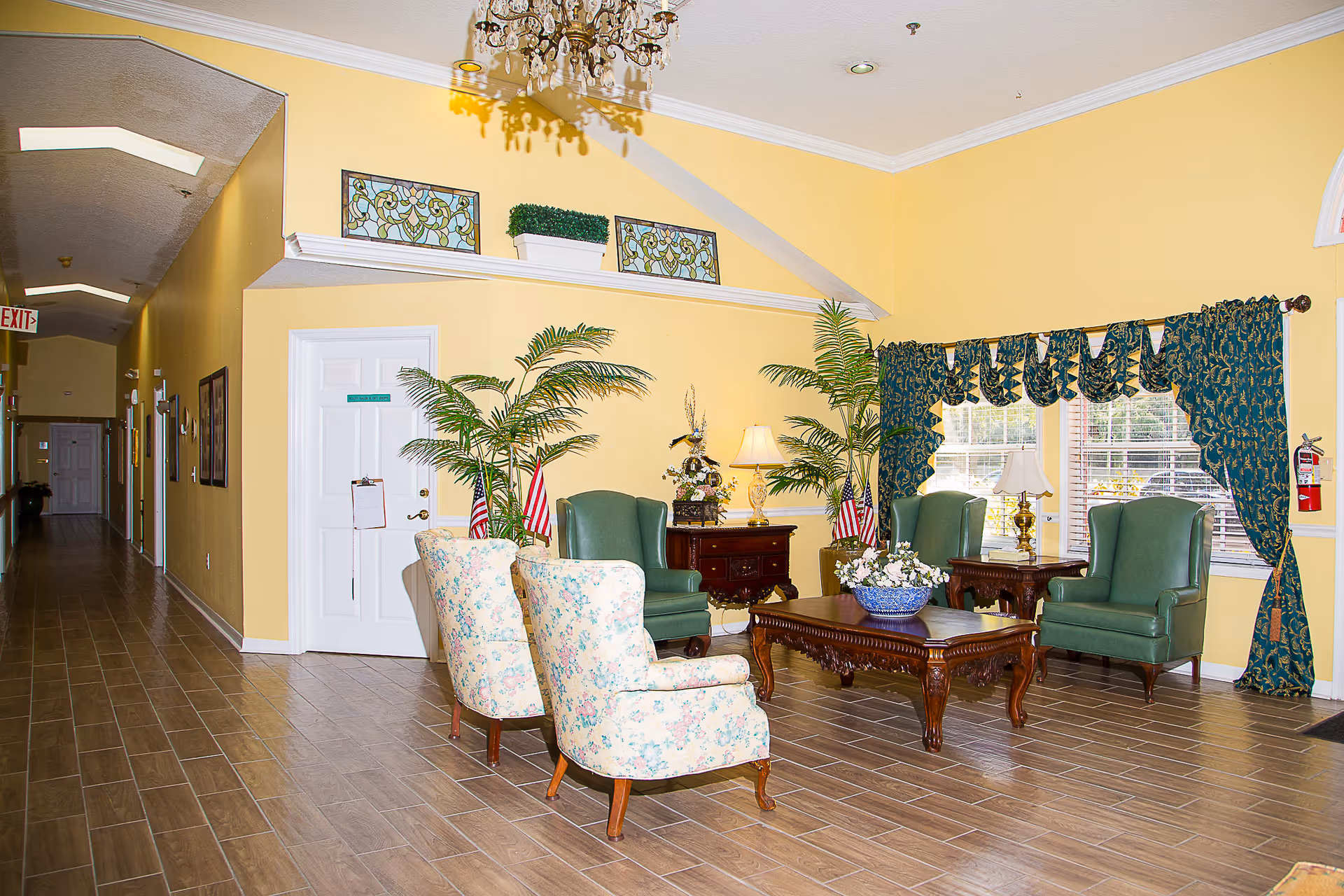 A bright and welcoming assisted living facility common area with yellow walls and wood-look tile flooring. The seating area includes two floral upholstered armchairs, two green leather wingback chairs, and wooden tables with decorative lamps and a flower arrangement. Large windows with dark green patterned curtains allow natural light to fill the room. There are potted plants and small American flags placed around the space. A hallway with multiple doors extends from the seating area, and a chandelier hangs from the ceiling.