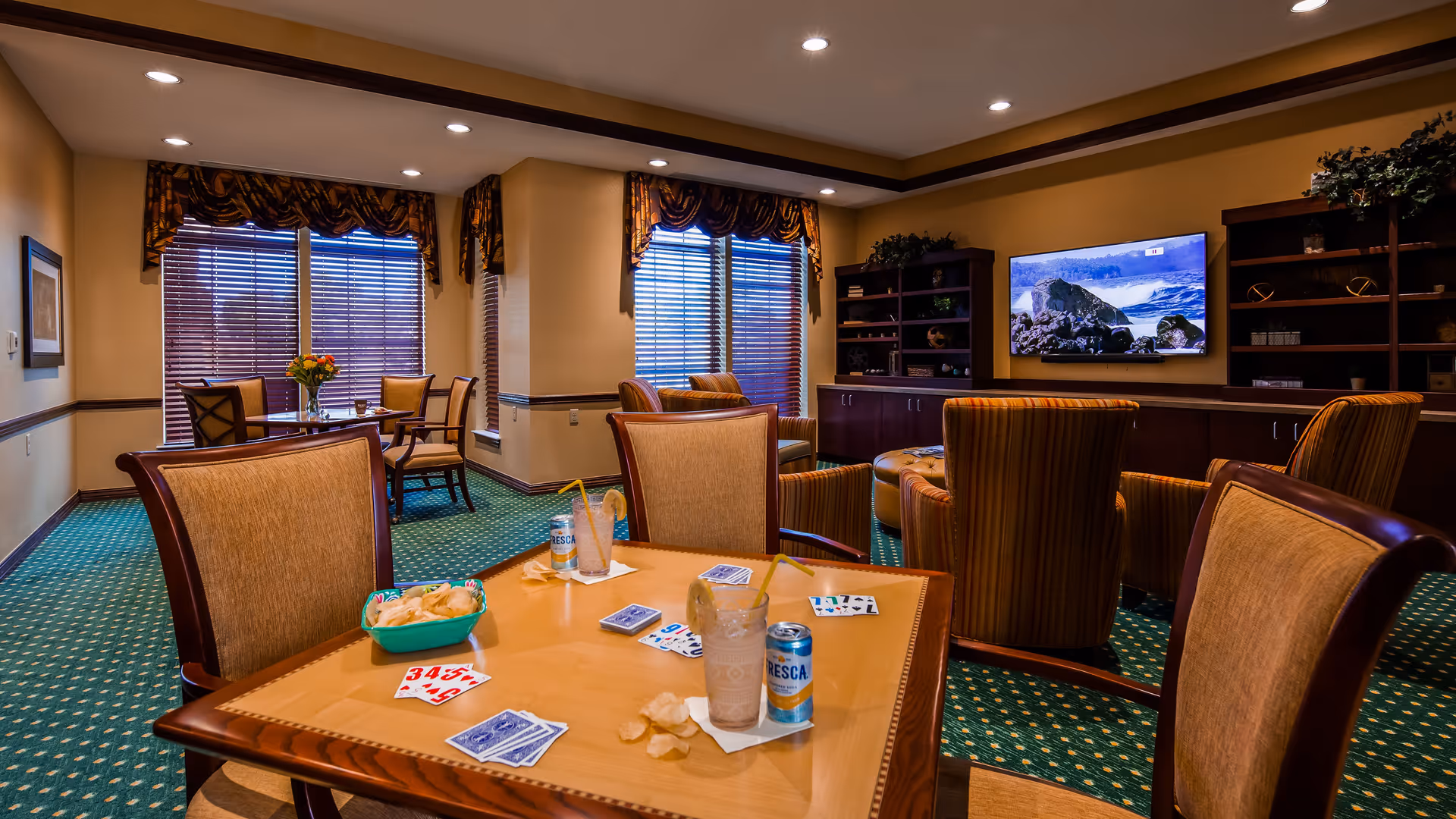 A cozy common area with a green carpet and beige walls featuring a table with playing cards, chips, and drinks. There are several upholstered chairs around the table and more seating in the background. A large flat-screen TV is mounted on the wall above a wooden cabinet with shelves, and windows with blinds and valances let in natural light.