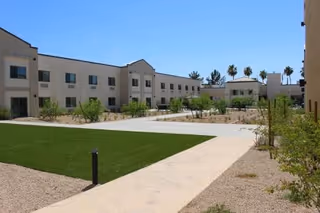 Outdoor courtyard area of BridgeWater Assisted Living - Deer Valley with a paved walkway, green lawn, desert landscaping, and a two-story beige building under a clear blue sky.
