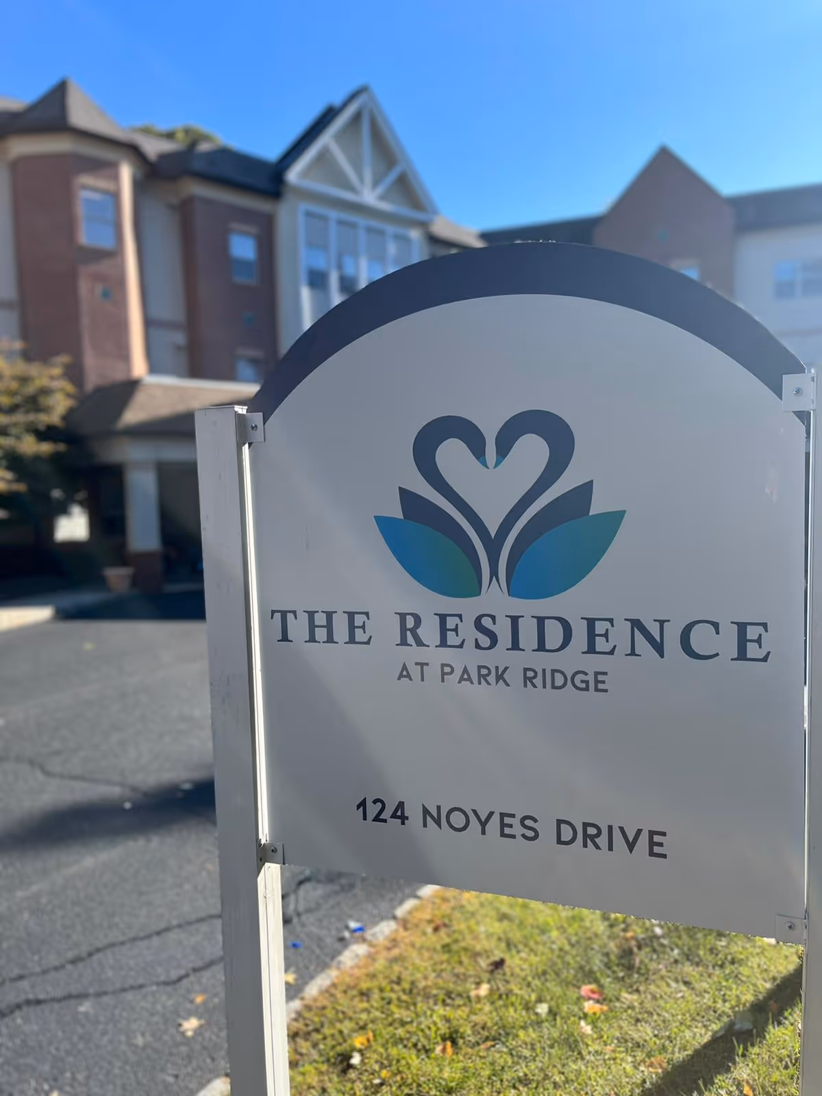 Outdoor view of a sign for The Residence at Park Ridge located at 124 Noyes Drive, with a building in the background under a clear blue sky.