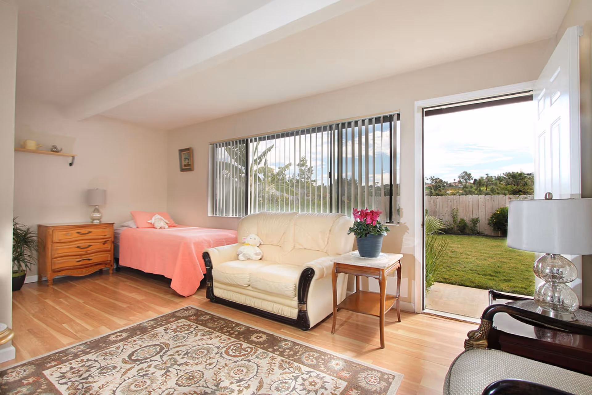 A bright room with light wooden flooring featuring a single bed with a pink bedspread, a wooden dresser with a lamp, a cream-colored loveseat with a stuffed animal, a small wooden side table with a potted plant, and a large window with vertical blinds. An open door leads to a green outdoor garden area with a wooden fence.