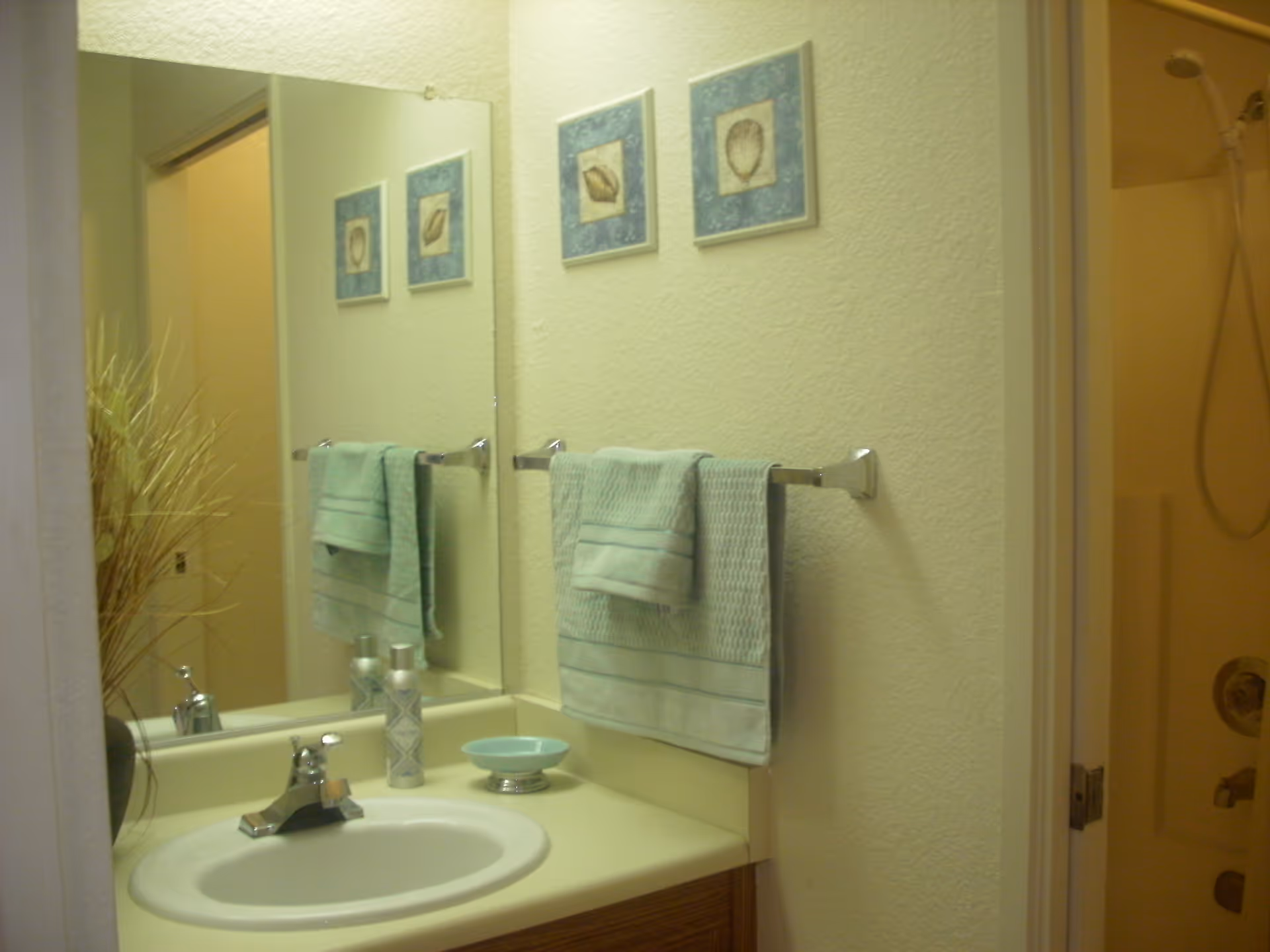 A bathroom sink with a faucet, a soap dish, and two decorative bottles on the countertop. Above the sink is a large mirror reflecting a towel rack with two folded green towels. On the wall above the towel rack are two framed pictures with seashell designs. To the right, there is a shower area with a showerhead and controls visible.