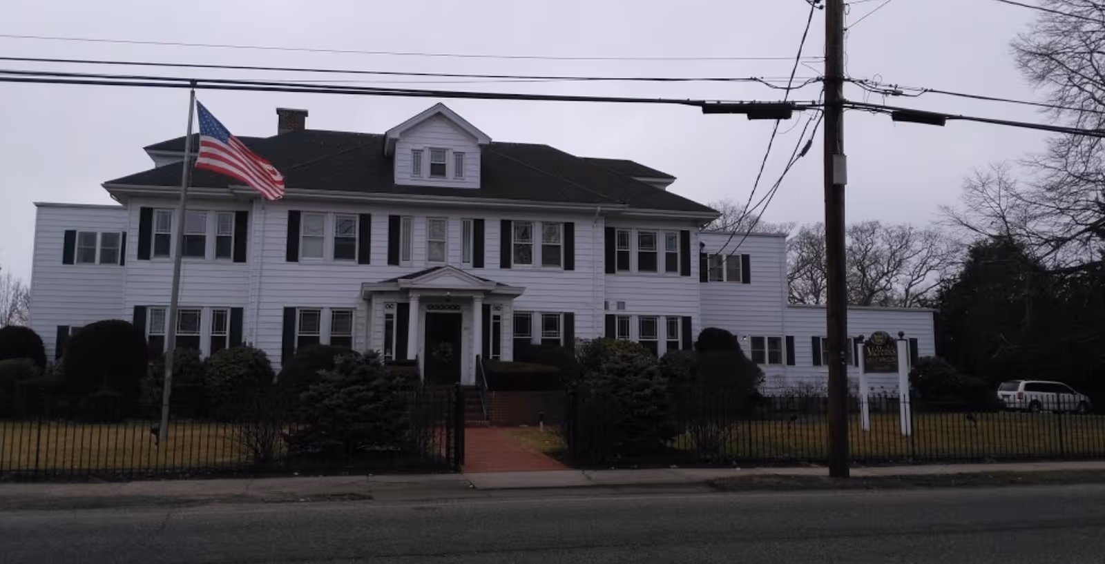 Front exterior view of a large white two-story building with black shutters, a dark roof, and a small porch entrance. An American flag is flying on a flagpole in the front yard, which is enclosed by a black metal fence. There is a sign on the right side of the yard indicating the facility name, McPeak's. Trees and bushes surround the building, and a utility pole with wires is visible in the foreground.