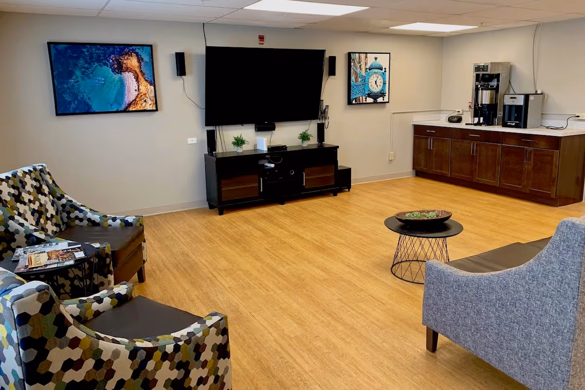 A cozy common area with patterned armchairs, a gray sofa, a small round black coffee table with decorative items, a large flat-screen TV mounted on the wall above a black media console, two framed pictures on the wall, and a countertop with a coffee maker and other appliances in the corner.