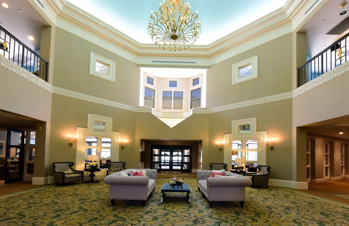 Spacious and elegant living room area with high ceilings, a large chandelier, two beige sofas with pink and blue cushions facing each other, a coffee table with flowers in the center, patterned green carpet, wall sconces, and windows near the ceiling allowing natural light.