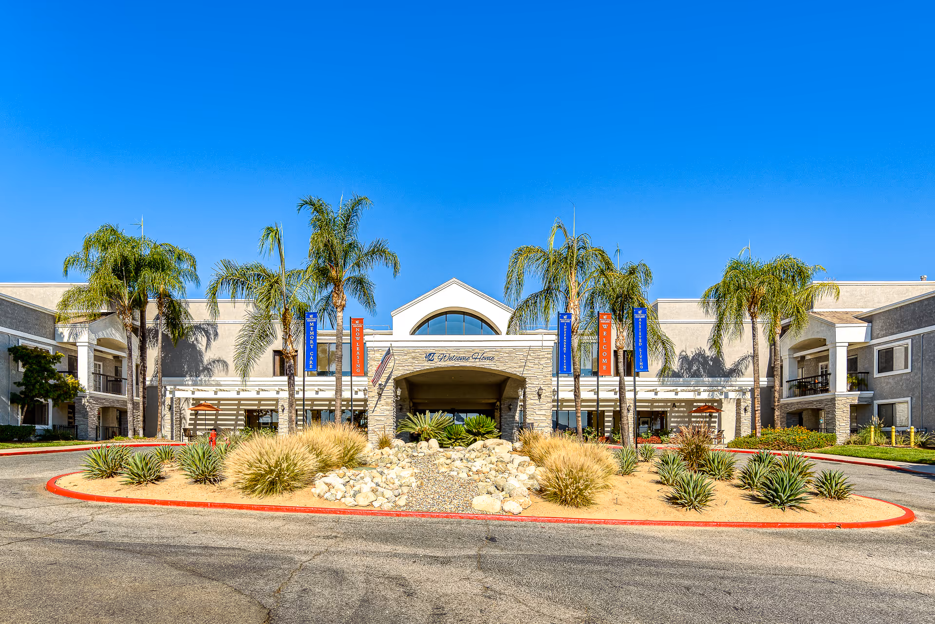 Front entrance of Menifee Senior Living with a circular driveway, landscaped rock island, palm trees and banners under a clear blue sky.