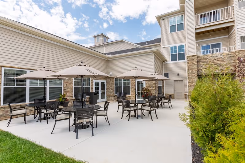 Outdoor patio area at StoryPoint Chesterton with several round tables and chairs under beige umbrellas, adjacent to a beige building with stone accents and multiple windows, under a partly cloudy sky.