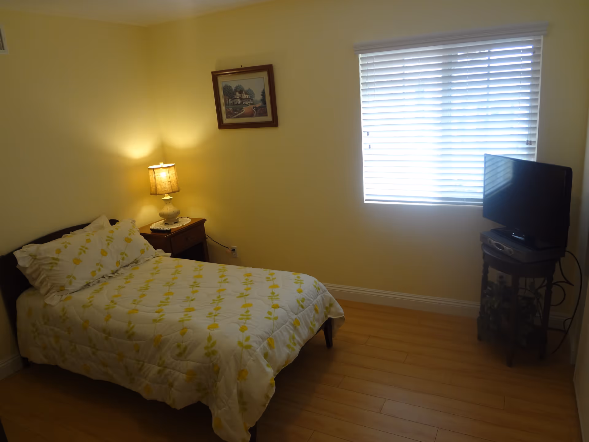 A small bedroom with a single bed covered in a white and yellow floral comforter. There is a wooden nightstand with a lamp beside the bed. A framed picture hangs on the wall above the nightstand. A window with white blinds is on the adjacent wall, and a small TV sits on a round wooden table in the corner. The floor is light wood, and the walls are painted a soft yellow.