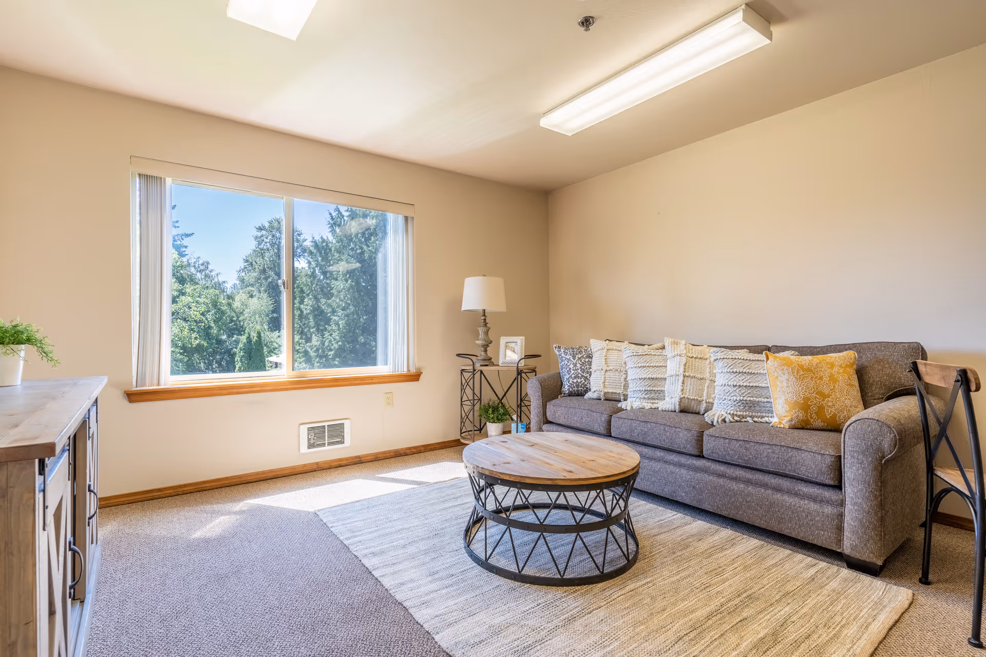 Sunny living room with a gray sofa, round wooden coffee table, large window, and neutral decor.