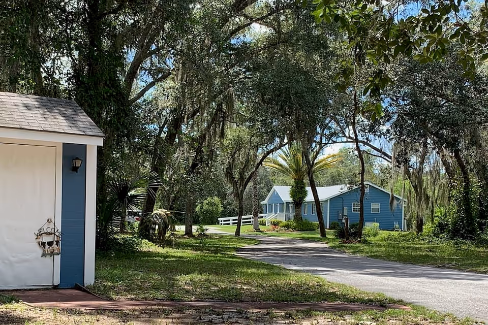 A peaceful outdoor scene at Silver Lake Village by Preferred Assisted Living featuring a blue building with a white porch in the background, surrounded by tall trees and greenery. In the foreground, there is a small blue and white shed with a 'Welcome' sign on the door, and a paved pathway leading towards the building.