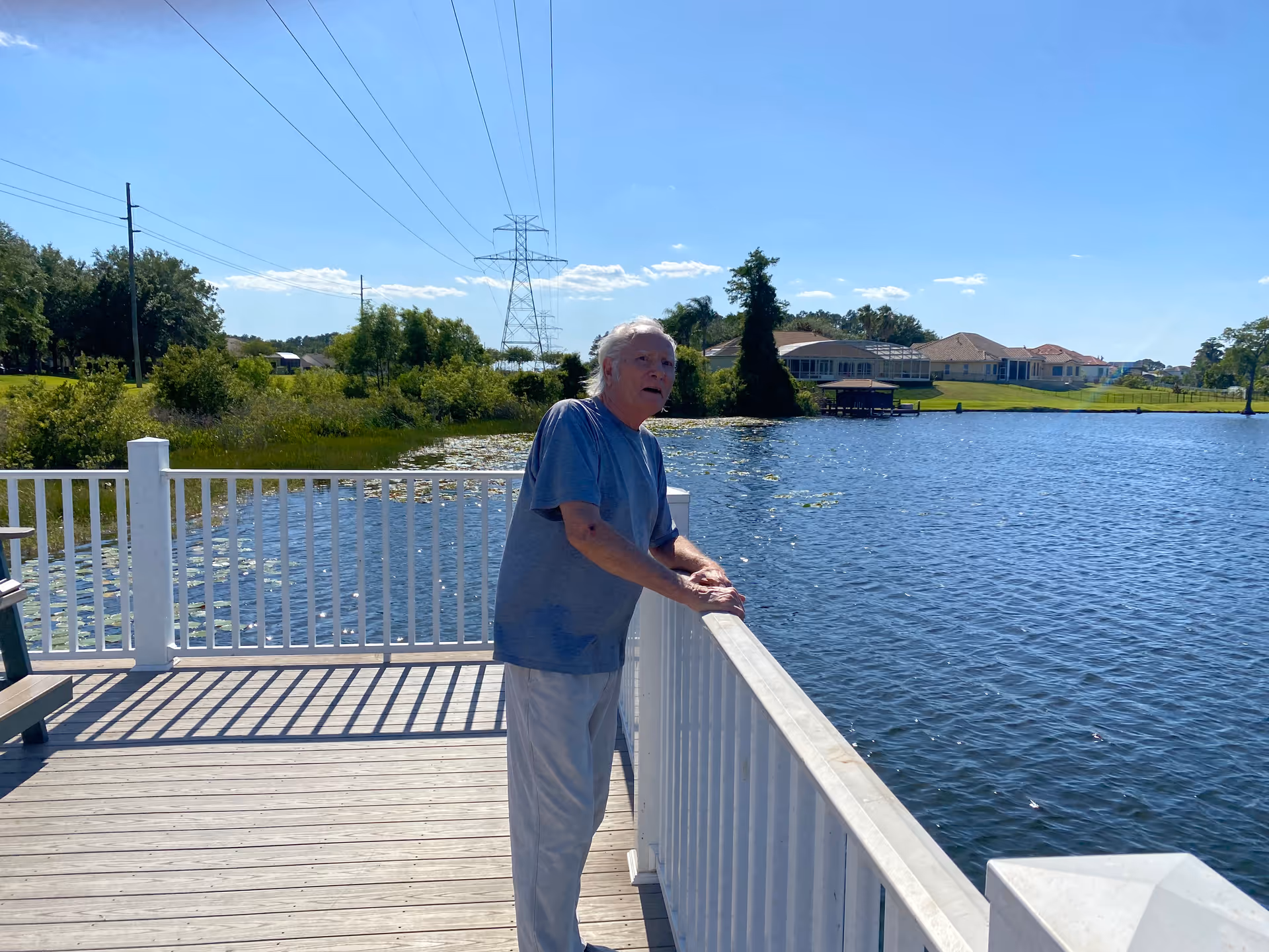 An elderly man wearing a blue shirt and light-colored pants stands on a wooden deck overlooking a large body of water with houses and greenery in the background under a clear blue sky.