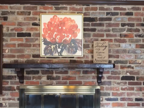 Brick fireplace with wooden mantle, red flower painting, and inspirational sign.