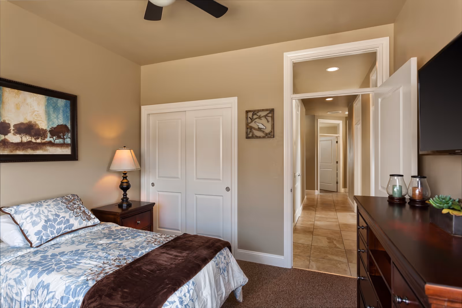 A cozy bedroom in an assisted living facility featuring a bed with floral bedding and a brown throw blanket, a wooden nightstand with a lamp, a framed artwork on the wall, a closet with white sliding doors, and a dresser with decorative items. The room opens into a hallway with tiled flooring.