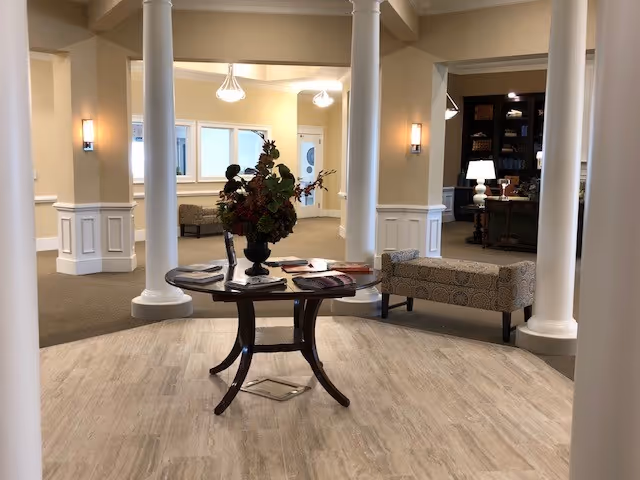 A warm interior lobby with a round table and floral centerpiece in the foreground, white columns, seating benches, and soft overhead lighting.