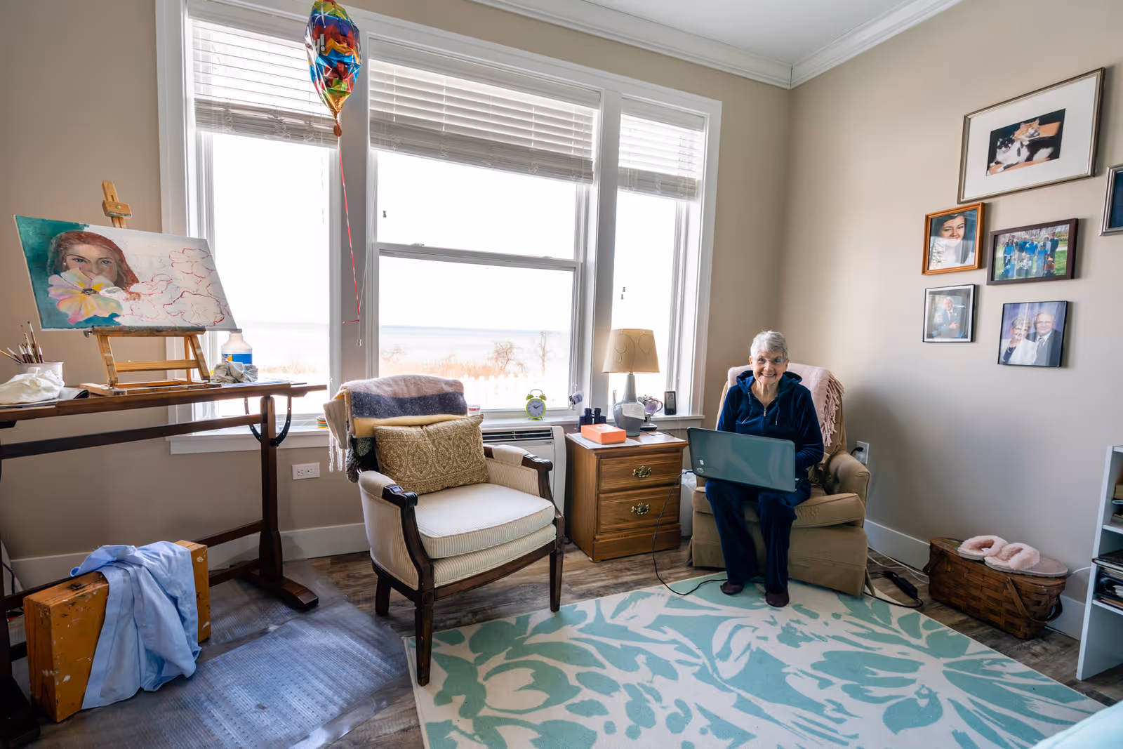 A senior woman sitting in a beige armchair using a laptop in a cozy room with large windows overlooking a lake. The room has a patterned blue and white rug, a wooden side table with a lamp, framed photos on the wall, an empty armchair with a cushion, and an easel with a painting of a woman holding a flower. A colorful balloon is tied near the window.