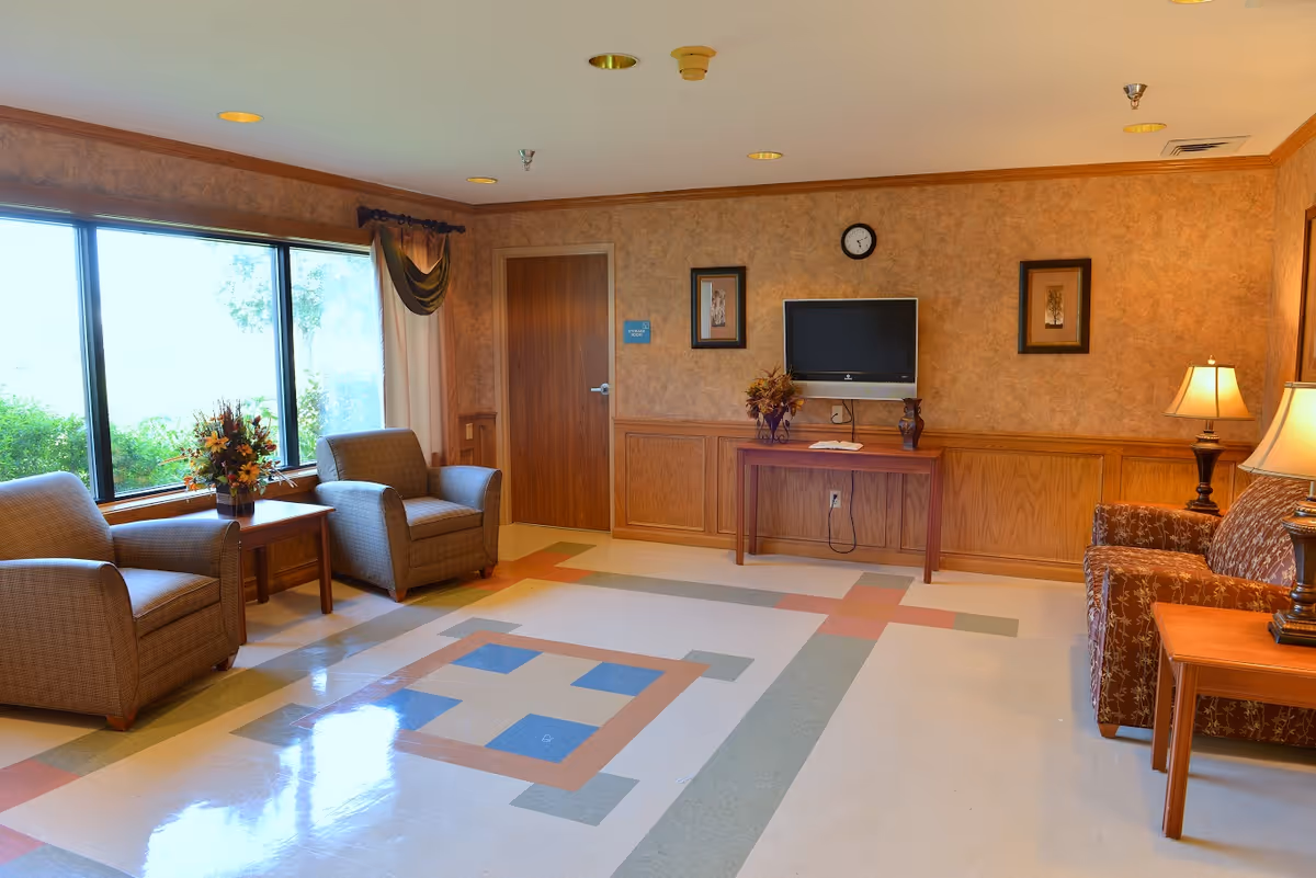 A cozy living room area in a senior living facility with two armchairs and a small table with a flower arrangement near a large window. Across the room is a wooden table with a flat-screen TV mounted above it, flanked by two framed pictures and a clock on the wall. There is also a patterned sofa with a side table and lamp, and the floor has a colorful geometric pattern.
