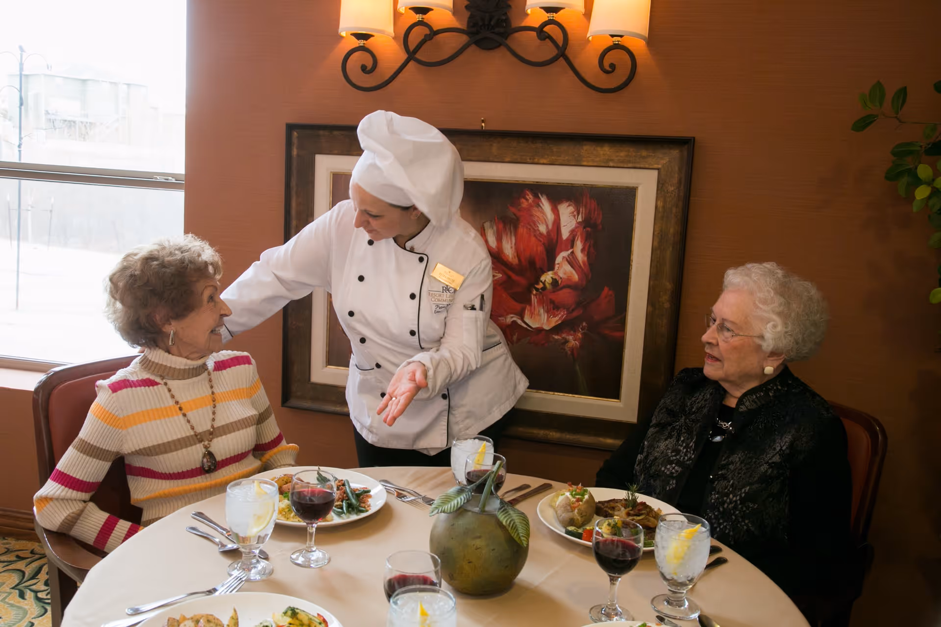 A chef in a white uniform and hat is interacting warmly with two elderly women seated at a round dining table set with plates of food, glasses of water with lemon, and glasses of red wine. The setting appears to be a cozy dining room with a large window, a framed floral painting on the wall, and warm lighting.