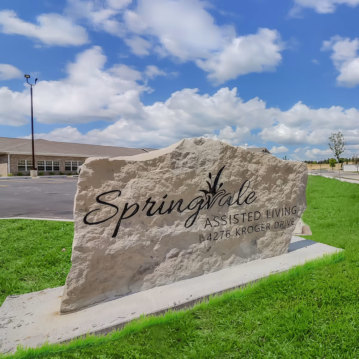 A large stone sign on a grassy area with the text 'Springvale Assisted Living 4276 Kroger Drive' engraved on it. In the background, there is a building and a parking lot under a partly cloudy blue sky.