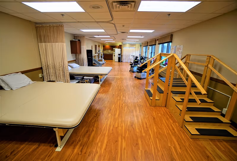 A rehabilitation therapy room with wooden flooring, two padded therapy tables with pillows on the left, exercise equipment including stationary bikes in the background, and a wooden staircase with handrails on the right side. The room is well-lit with ceiling lights and has large windows along the right wall.
