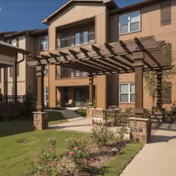 Outdoor patio area at a senior living facility with a wooden pergola, seating, and landscaped garden beds in front of a multi-story building with balconies and windows.