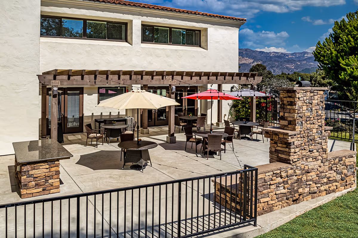 Outdoor patio at a senior living facility with round tables and umbrellas under a wooden pergola, stone pillars, and mountains in the background.