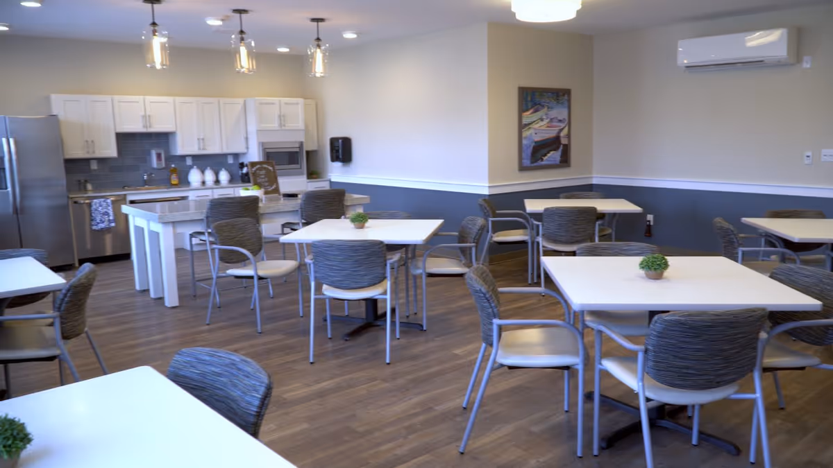 A clean and modern dining area with several square tables and cushioned chairs arranged neatly. The room features a kitchen area with white cabinets, a stainless steel refrigerator, a microwave, and a dishwasher. Pendant lights hang over a kitchen island, and there is a framed painting on the wall. The floor is wooden, and small potted plants are placed on the tables.